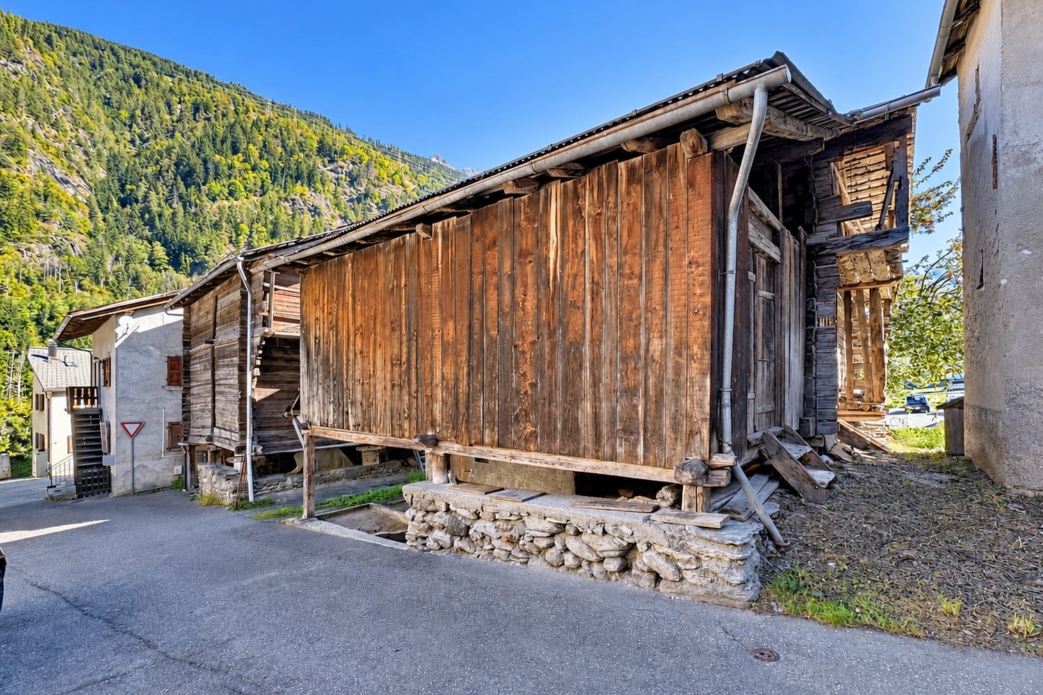 L'annexe en planches et le pied de pierre du raccard, avec le versant boisé du Val de Bagnes en arrière-plan.